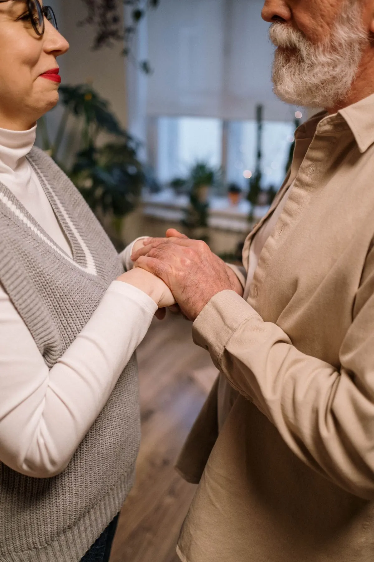 An older couple faces one another and holds hands in their living room.