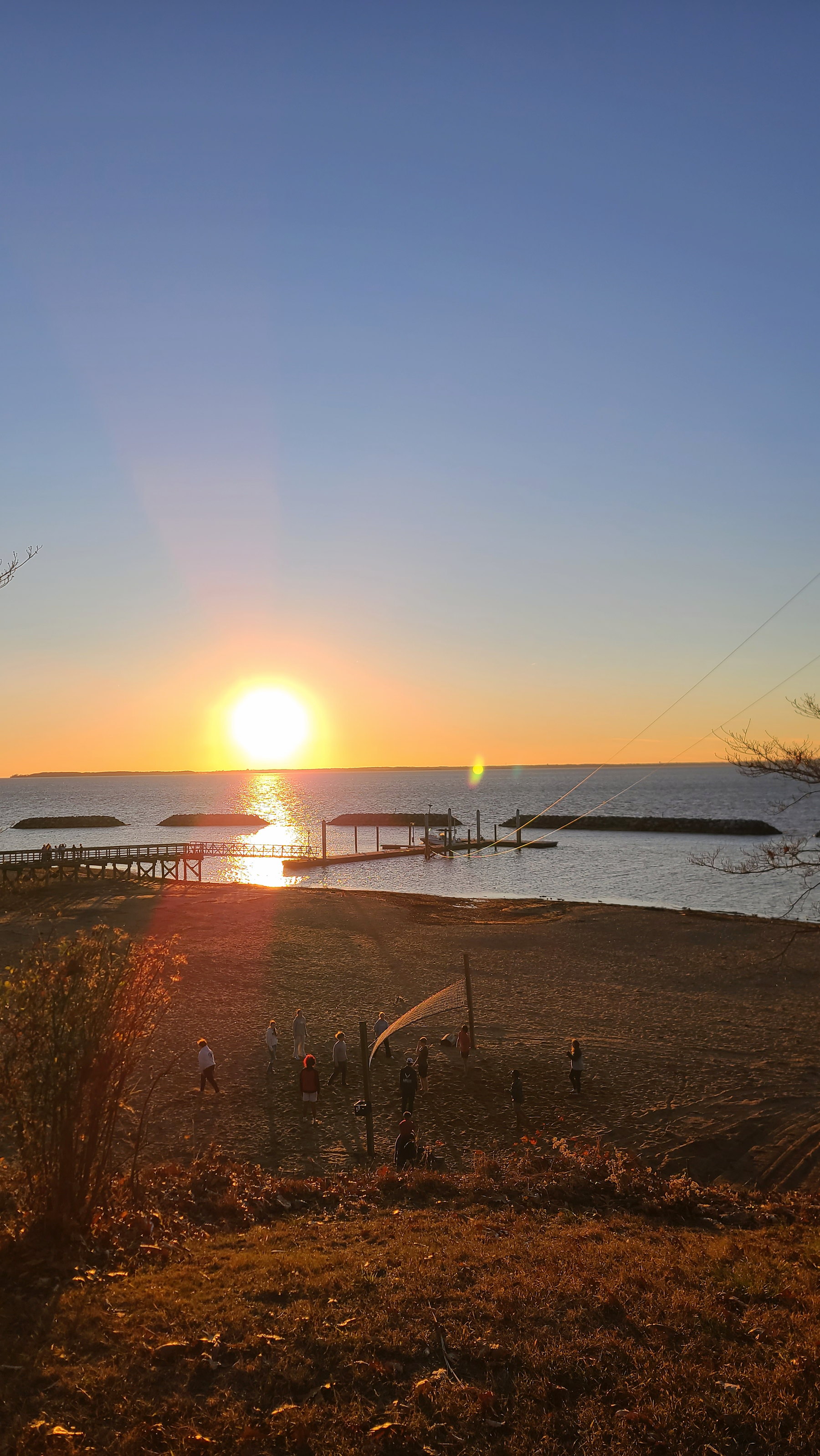 Sunset over a waterfront volleyball court where small groups gather near the shore.